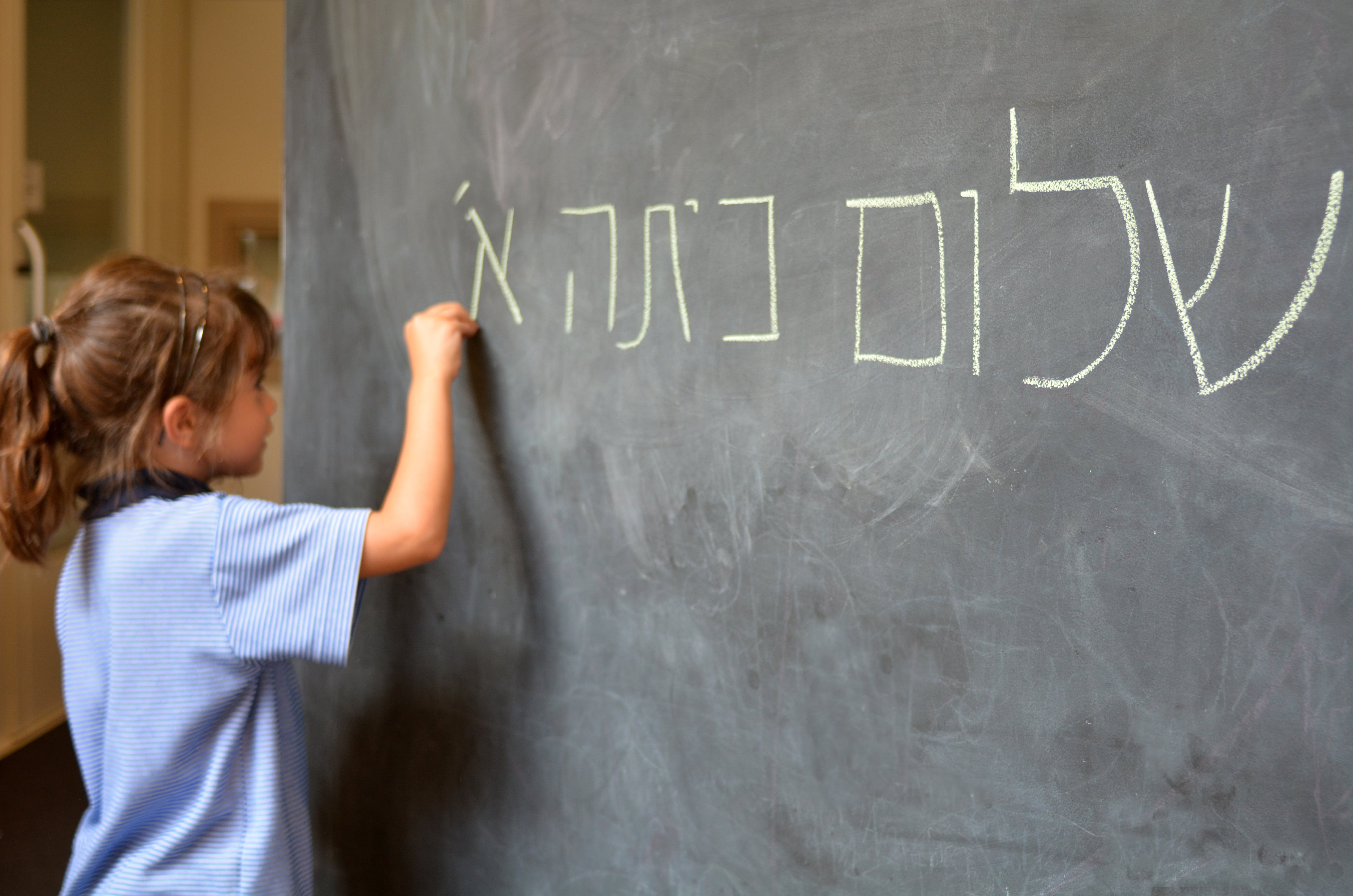 Photo d'une petite fille blonde tenant un bout de craie pour écrire en hébreux sur un tableau noire
